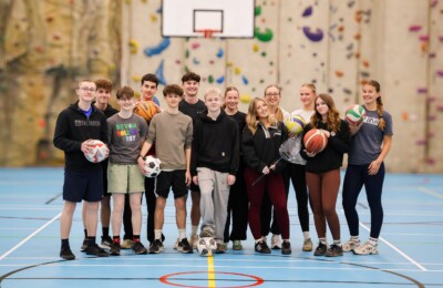 group of students stood in the sports hall holding a variety of sports balls